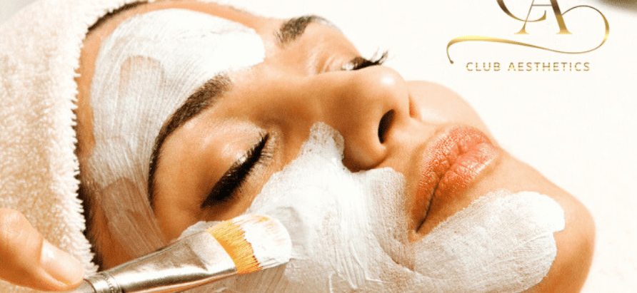 Woman receiving a facial mask with a brush, eyes closed, relaxed. Logo "Club Aesthetics" in the corner, spa setting, soft lighting.
