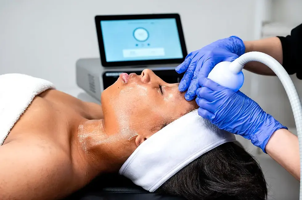 Person receives facial treatment with a blue-handled device. They have a headband and towel, and a screen is visible in the background.