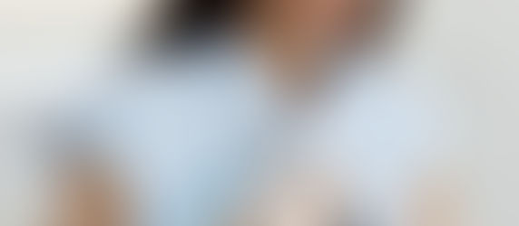 A woman in blue scrubs performs a facial treatment on a reclining person in a headband. Bright setting with a calm, focused mood.