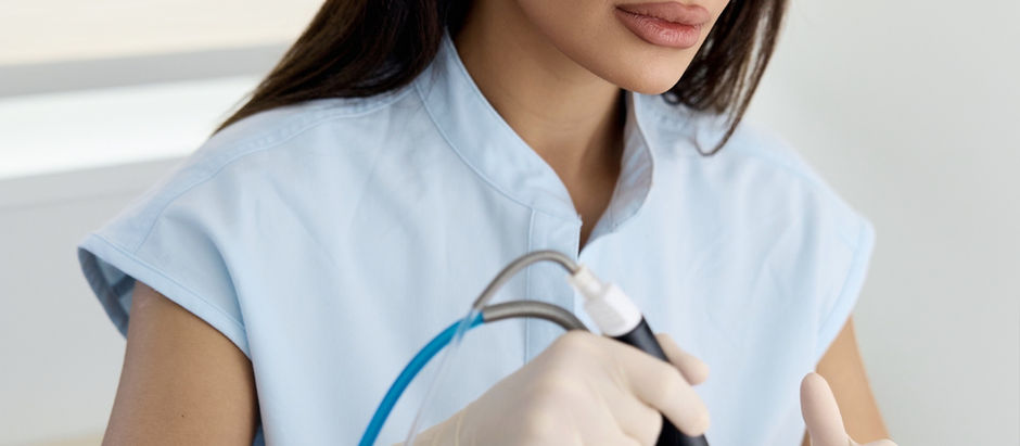 A woman in blue scrubs performs a facial treatment on a reclining person in a headband. Bright setting with a calm, focused mood.
