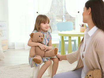 Child psychology session featuring a young girl holding a teddy bear, engaging in play-based therapy with a supportive clinic