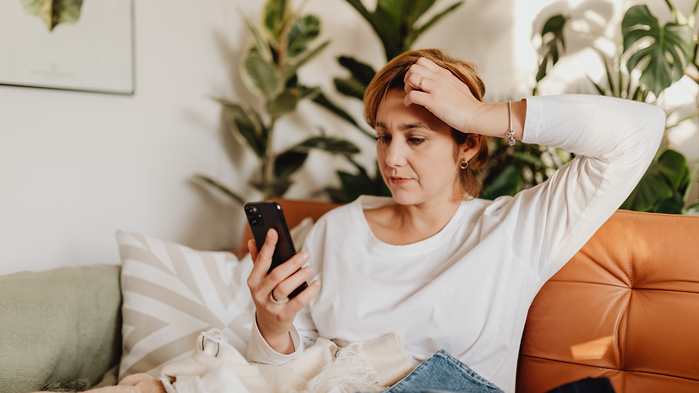 Woman in white shirt, sitting on an orange couch, looks at phone with a stressed expression. Green plants and patterned pillows in the background.