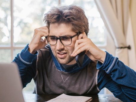 Man with glasses sits at table, talking on phone, looking frustrated. Open laptop on table. Bright room with curtains in background.
