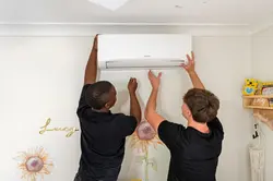 Two professional technicians carefully install a white split system air conditioner on a wall.