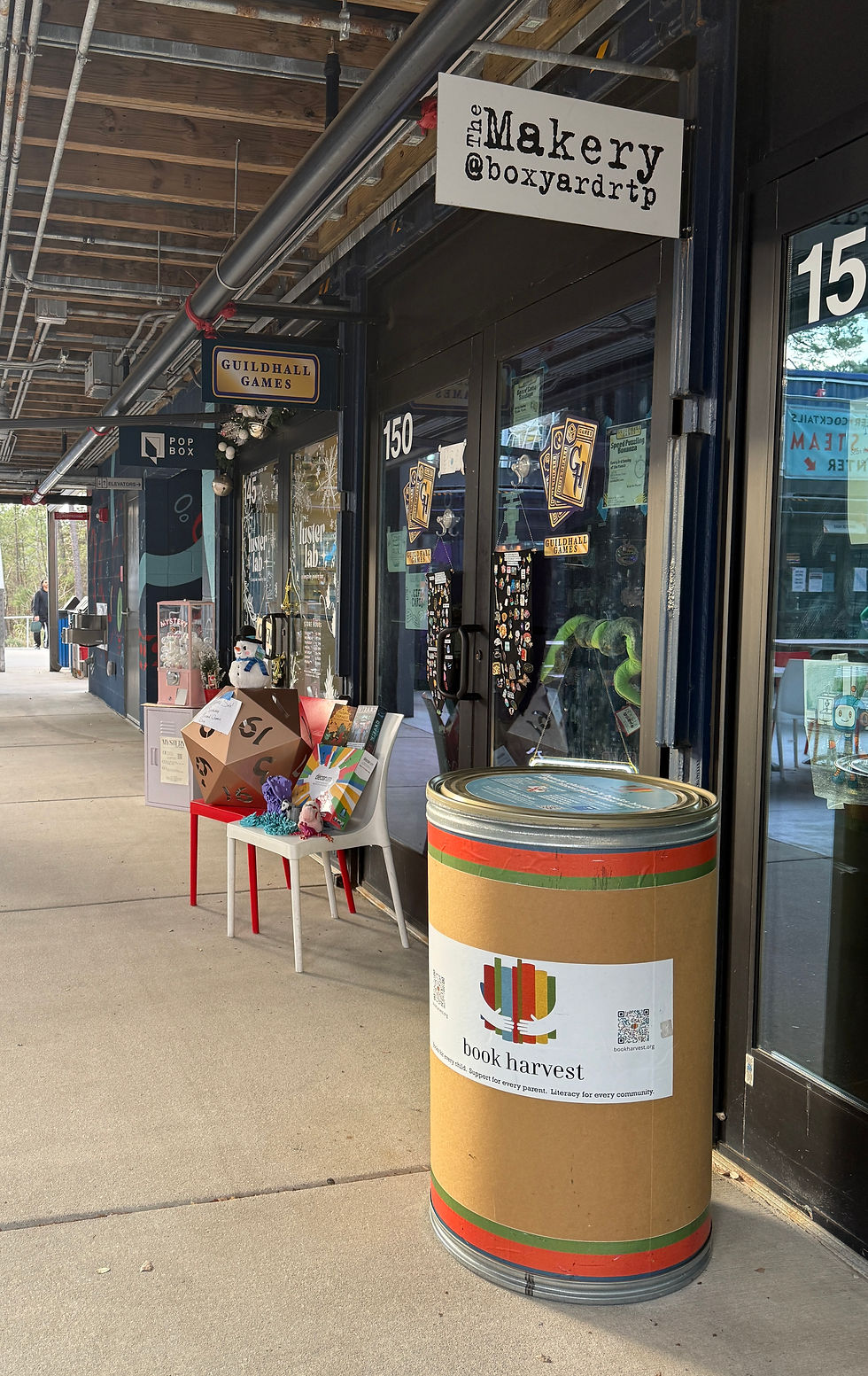 A book drive bin outside of The Makery at Boxyard RTP. It is a cylindrical bin with the Book Harvest logo in the middle.