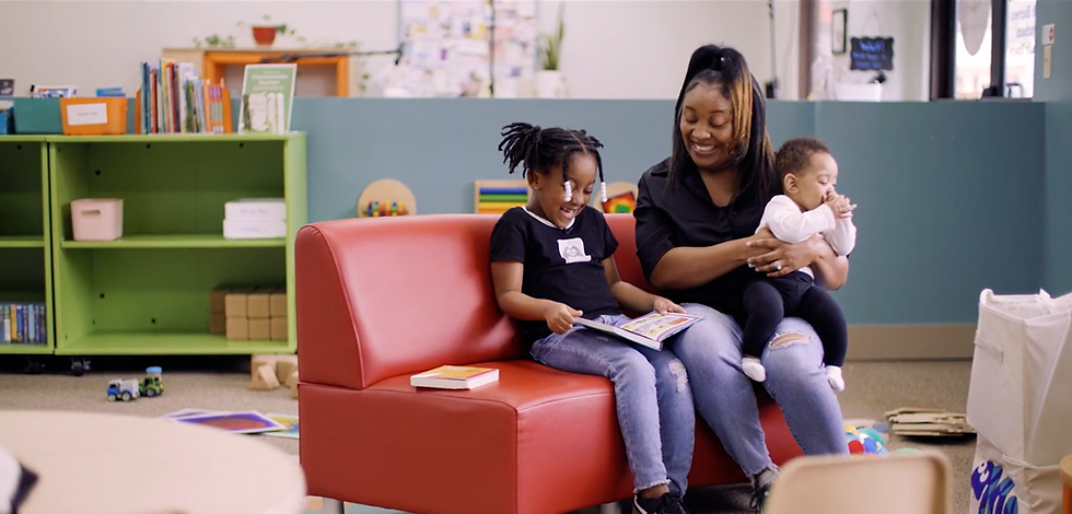 A mother reads with her 6-year-old daughter while holding her baby.