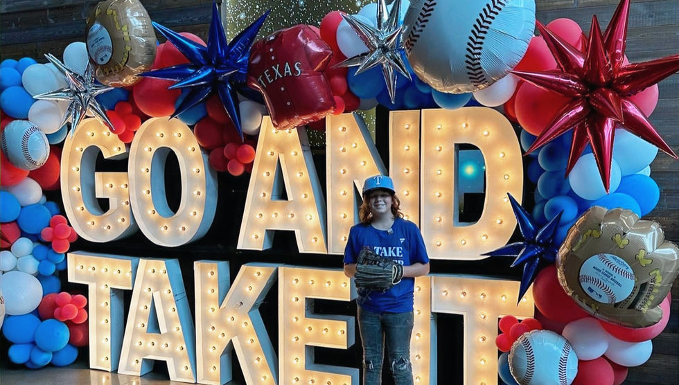 McKennon at Globe Life Field before his Texas Rangers Playoff Experience with a big sign that says "Go And Take It" in the background with red white and blue balloons. 