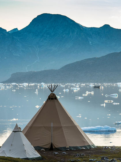 Tipi tents set against a dramatic backdrop of a fjord, icebergs, and rugged mountains in Greenland, offering unique lodging experiences.