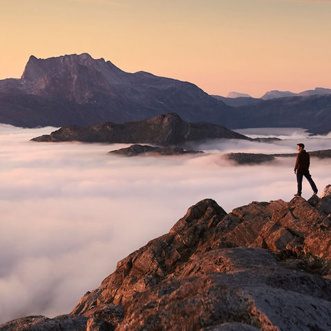 Hiker on a premier Greenland hiking tour, standing on a mountain peak at sunrise with views of a sea of clouds and rugged landscapes, capturing a unique nature exploration experience.
