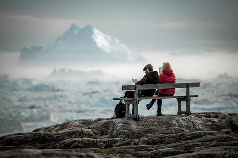 Two travelers enjoying a boutique Greenland experience, reading on a bench with views of icebergs in the Ilulissat Icefjord, capturing the tranquility and unique beauty of Arctic landscapes.