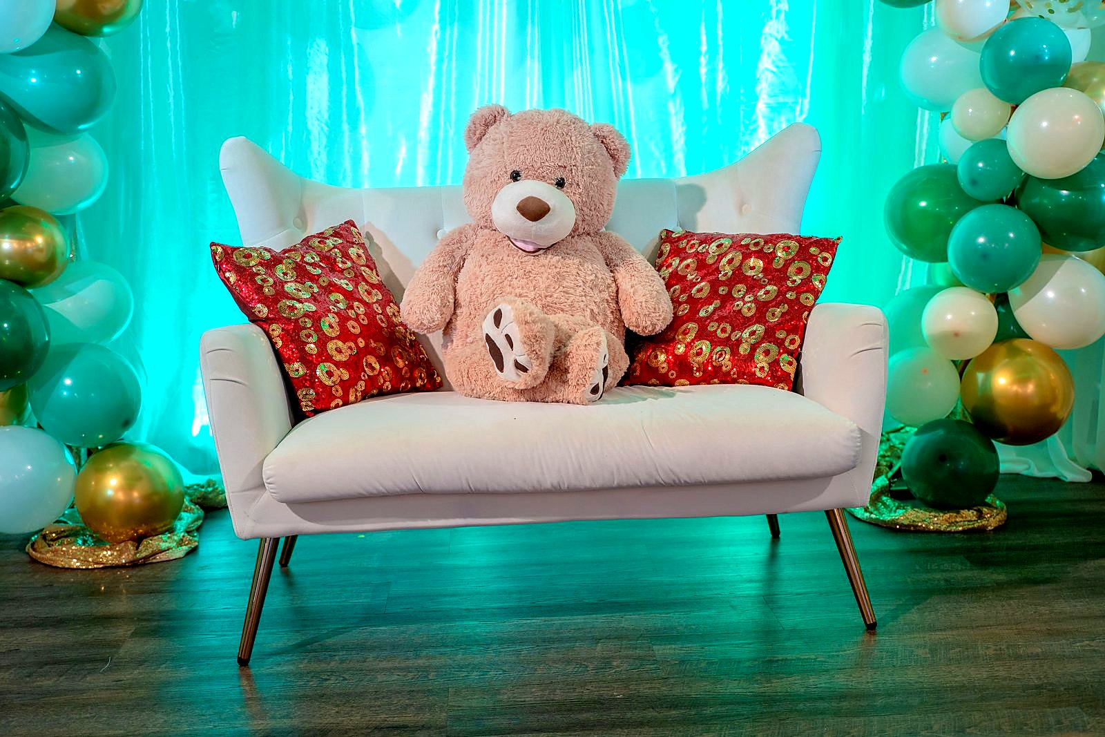 Teddy bear on a white loveseat with sequin pillows, framed by green, white and gold balloon garlands