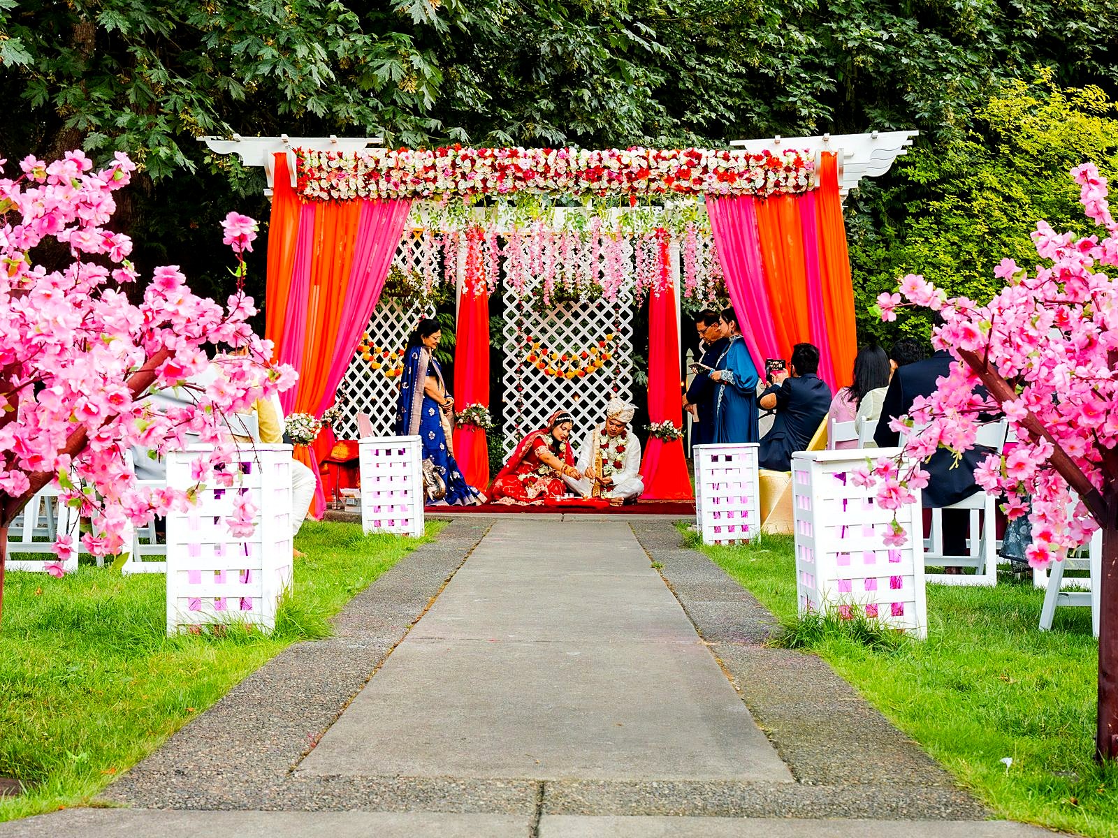 Outdoor wedding ceremony under the pergola – floral mandap, cherry-blossom trees lining the aisle and guests on the lawn