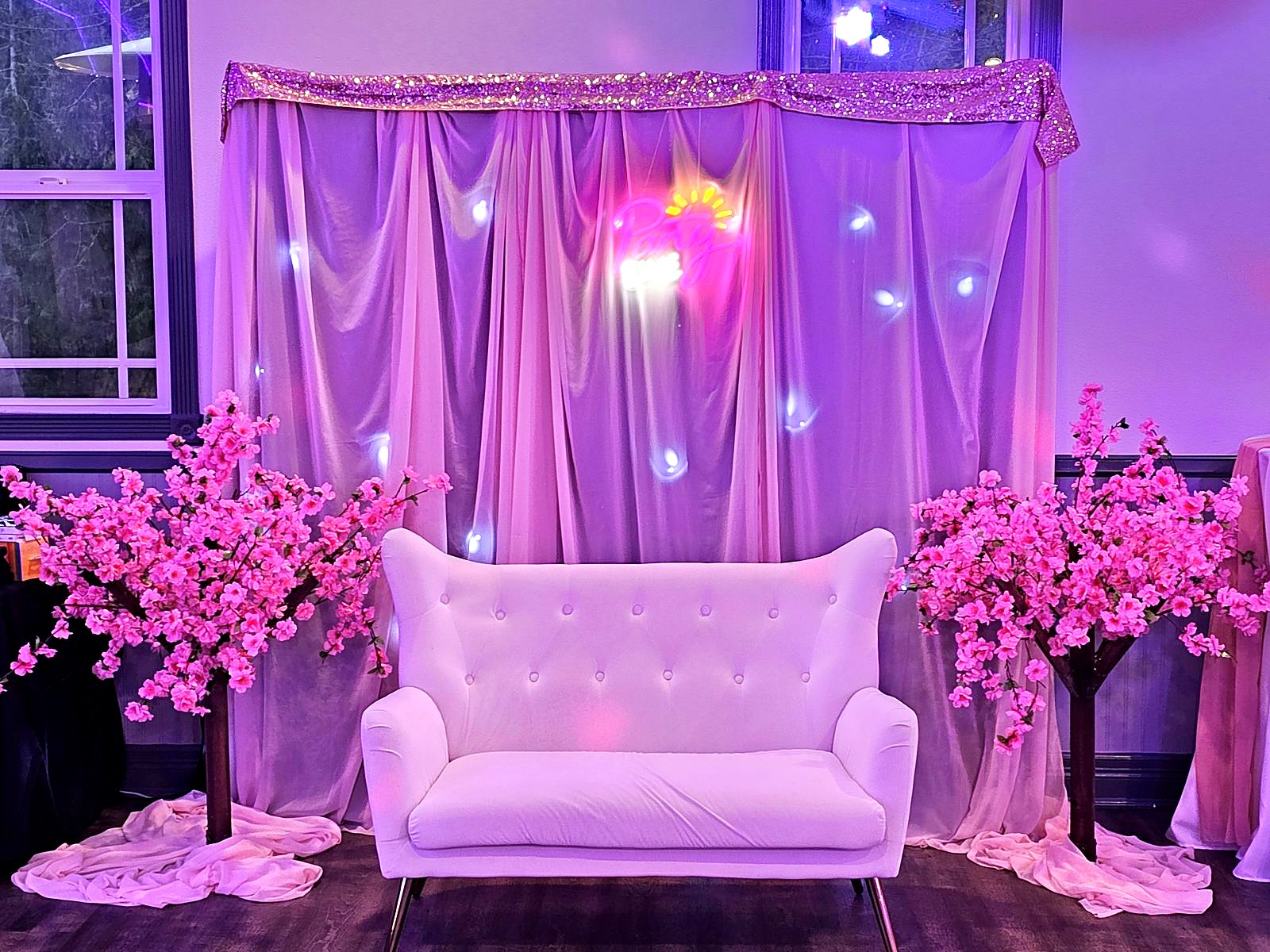White tufted loveseat between pink cherry-blossom trees with a neon "Party Time" sign
