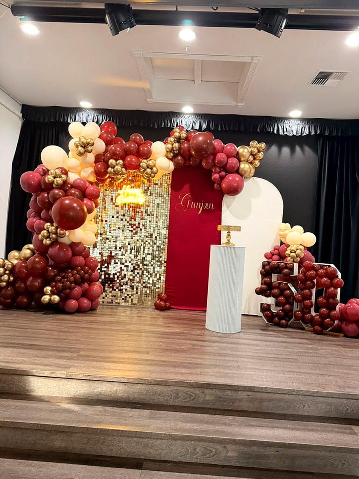 Burgundy and gold 50th birthday stage – gold shimmer wall, “Happy Birthday” neon and balloon-filled 50 marquee numbers