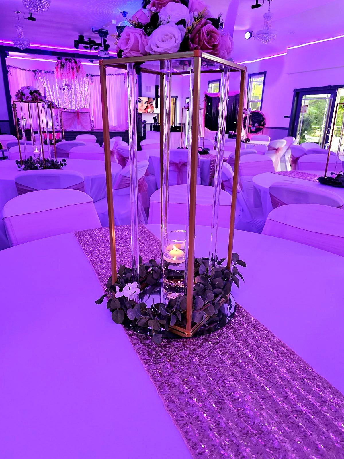 Tall gold-and-acrylic stand with blush roses and a floating candle on a sequin runner, in the pink-lit hall