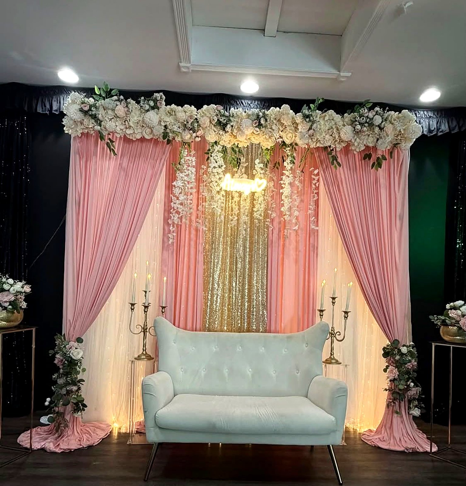 Blush and ivory draped backdrop with a cream floral garland, gold sequin panel, "Always" neon sign and a white loveseat between candelabras