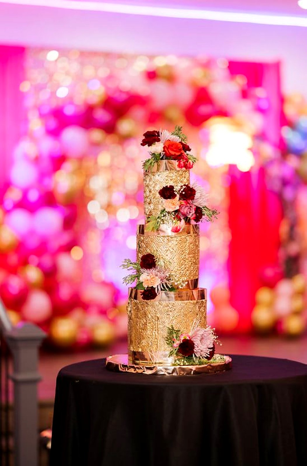 Three-tier gold leaf cake with burgundy and blush flowers in front of a pink and gold balloon wall