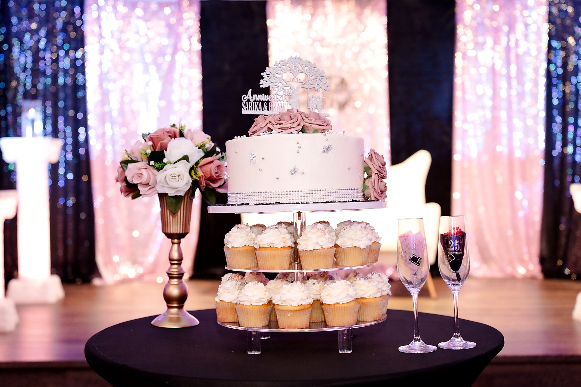25th anniversary cake and cupcake tower in front of a blush and black sequin backdrop, with engraved champagne flutes