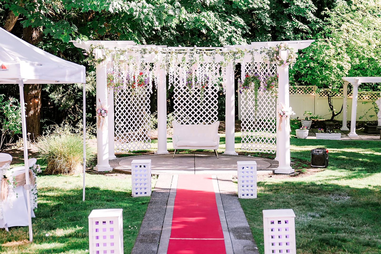 Wisteria-draped pergola with a white loveseat and red carpet aisle lined with lanterns