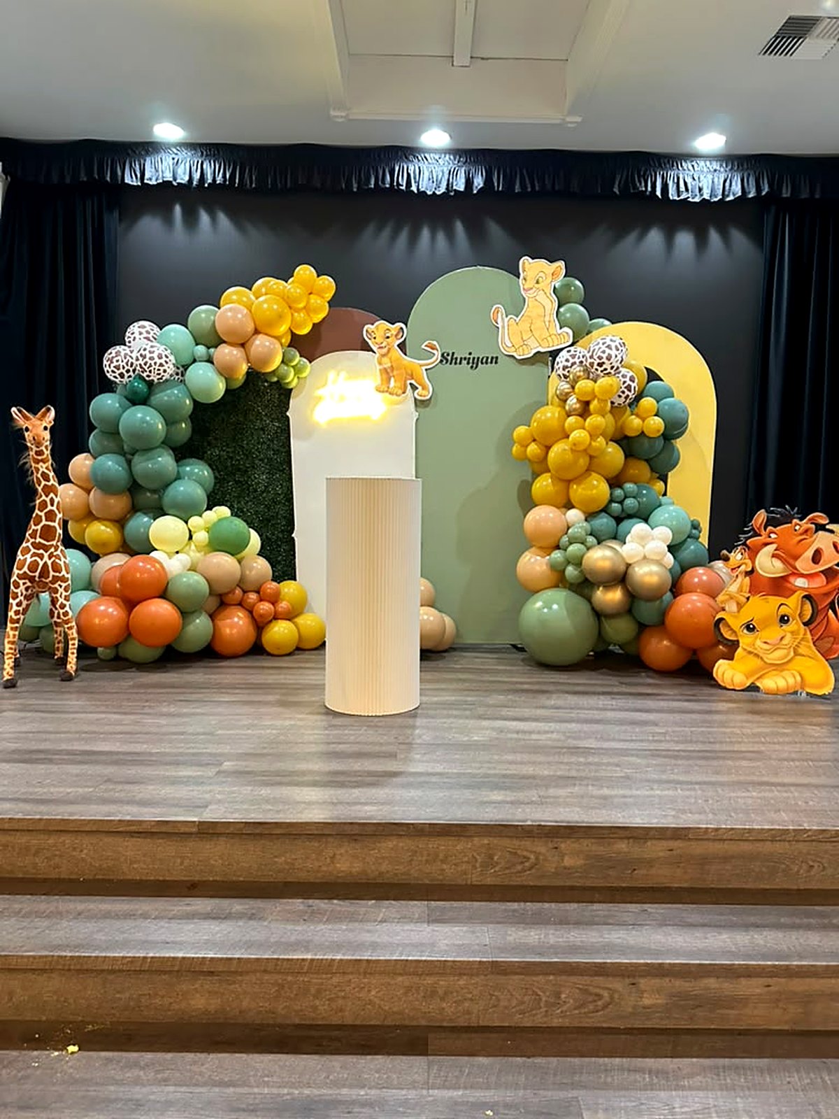 Jungle safari birthday stage – sage, mustard and terracotta balloon arches, greenery wall and a giraffe