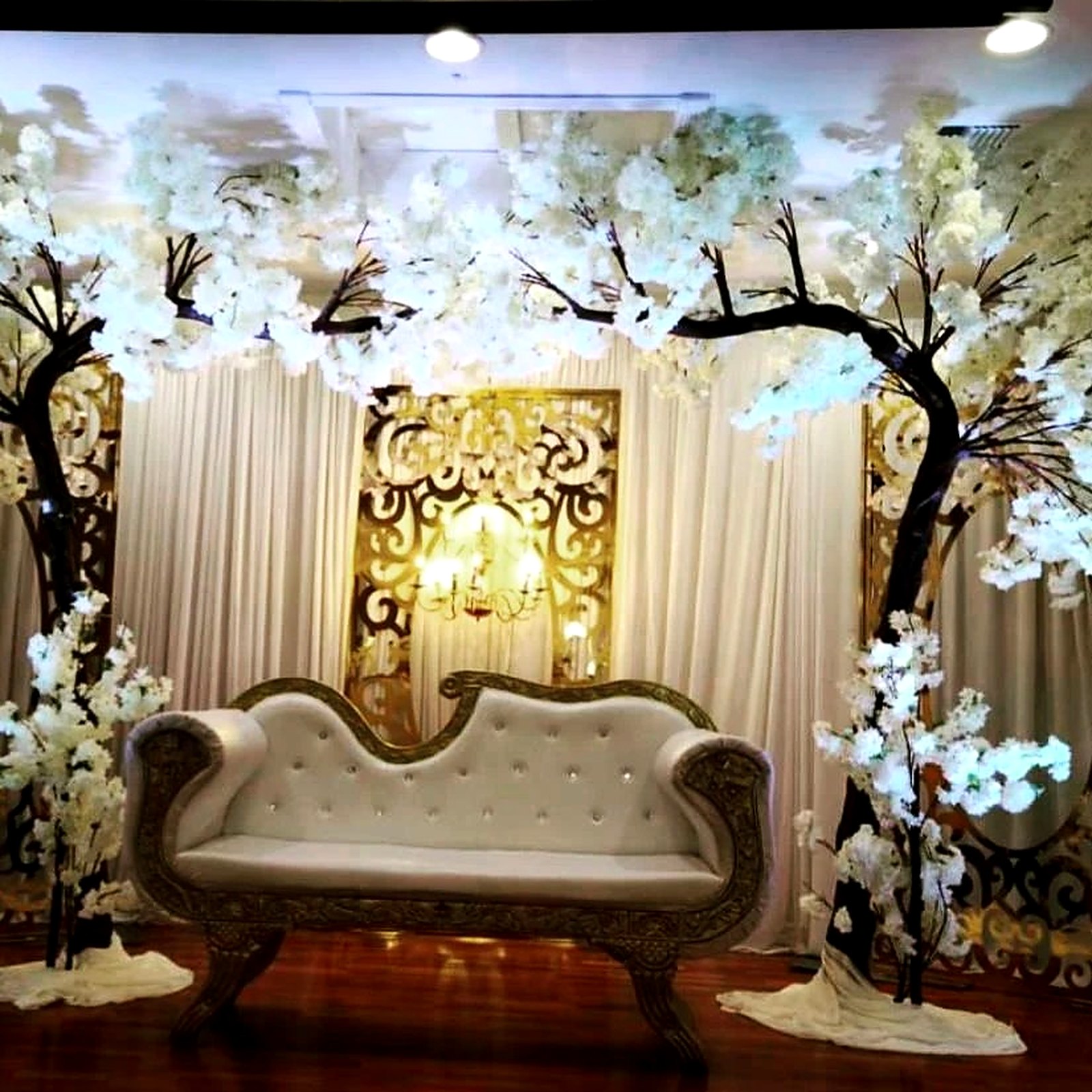 Sweetheart stage under white cherry-blossom trees – ivory and gold tufted settee in front of gold filigree panels, a crystal chandelier and white drapes