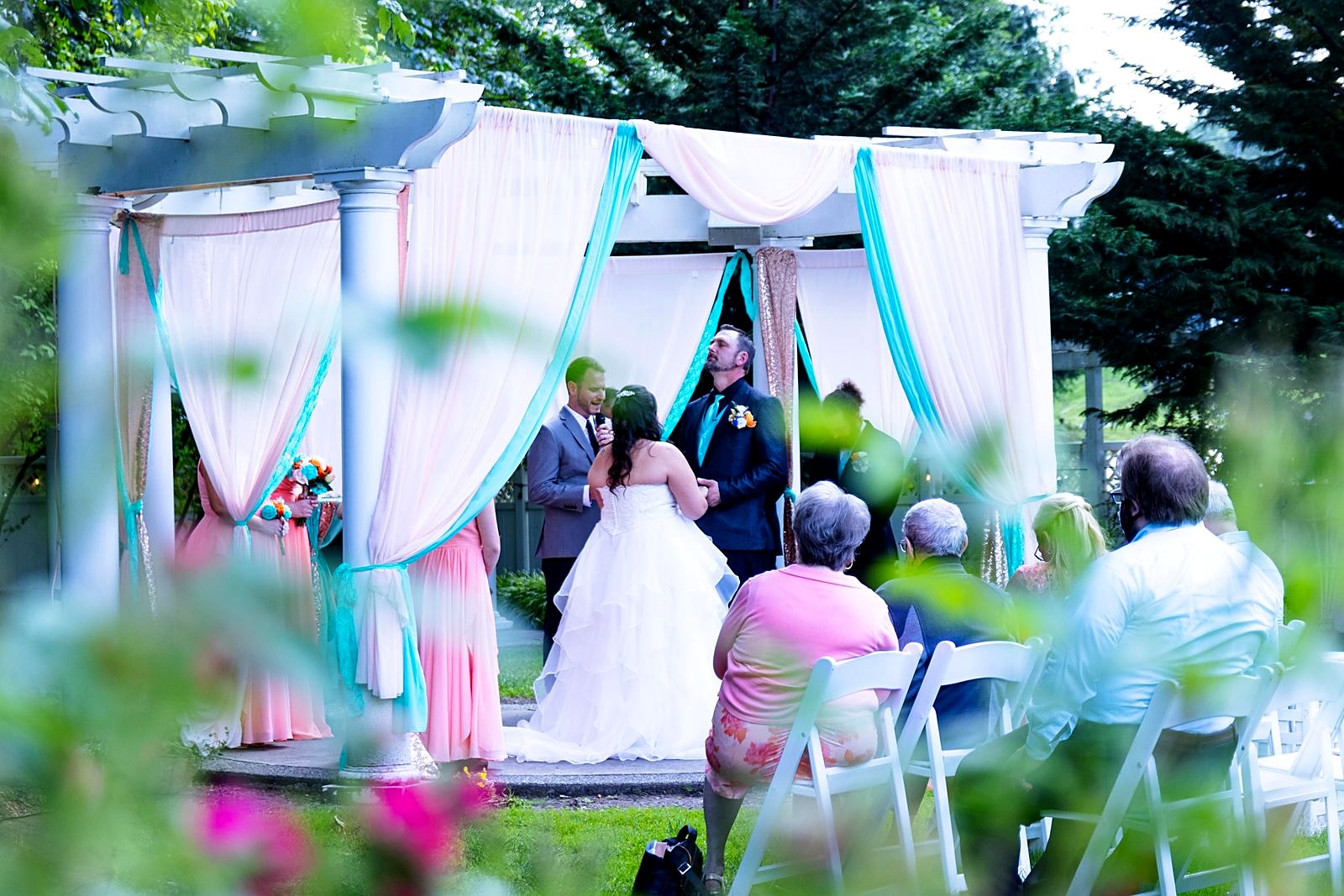 Couple exchanging vows under the blush and teal pergola, seen through the garden roses