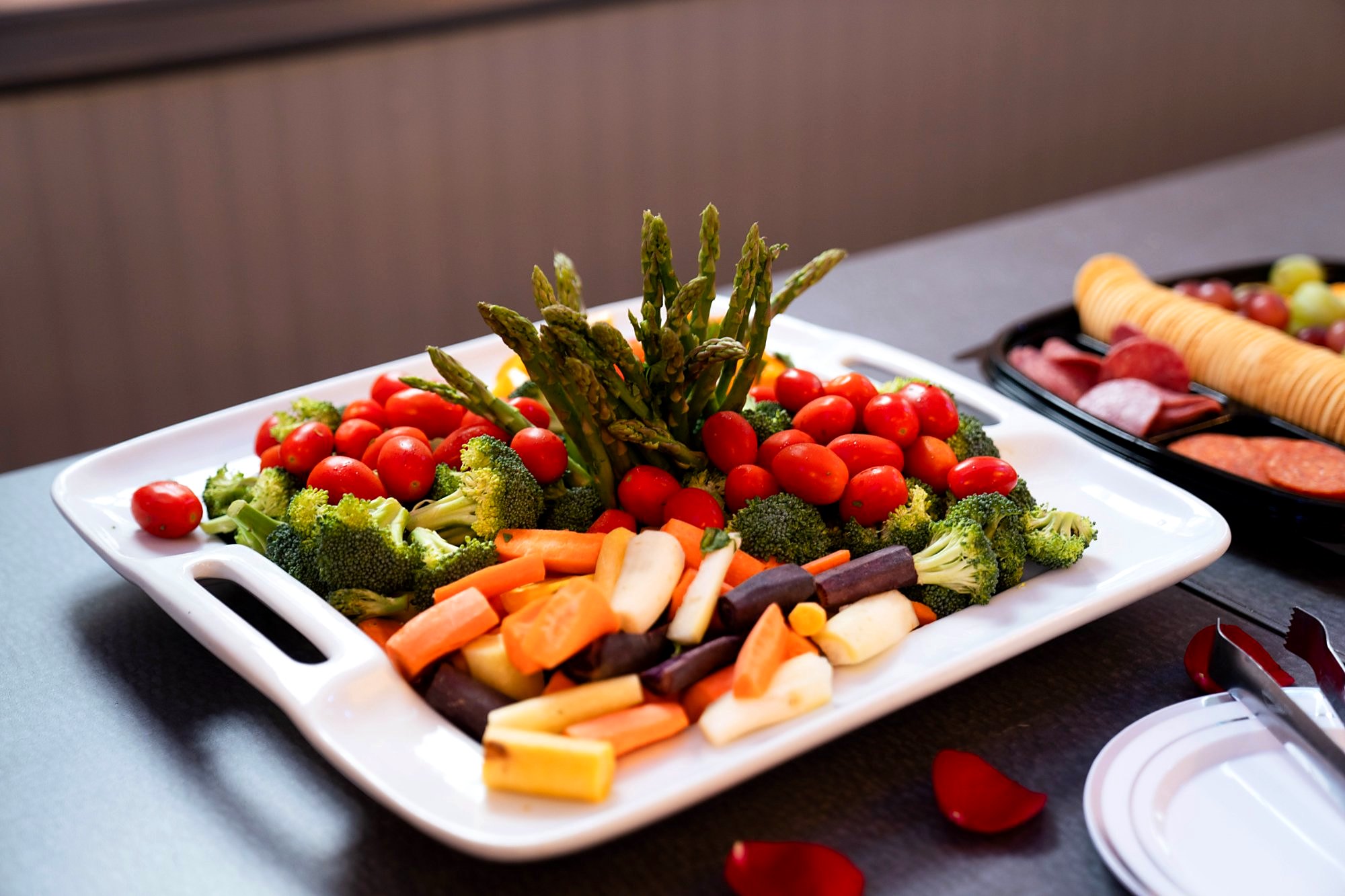 Fresh crudité platter – asparagus, cherry tomatoes, broccoli and rainbow carrots – beside a cheese, salami and cracker tray