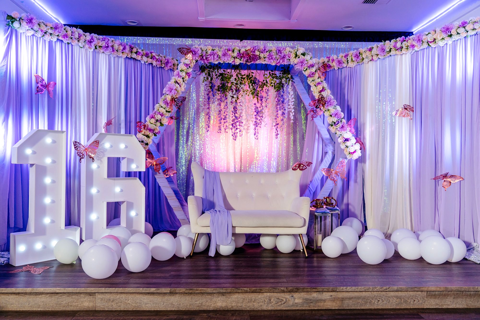 Sweet 16 stage – light-up 16 marquee numbers, lavender floral hexagon arch, white loveseat, butterflies and white balloons