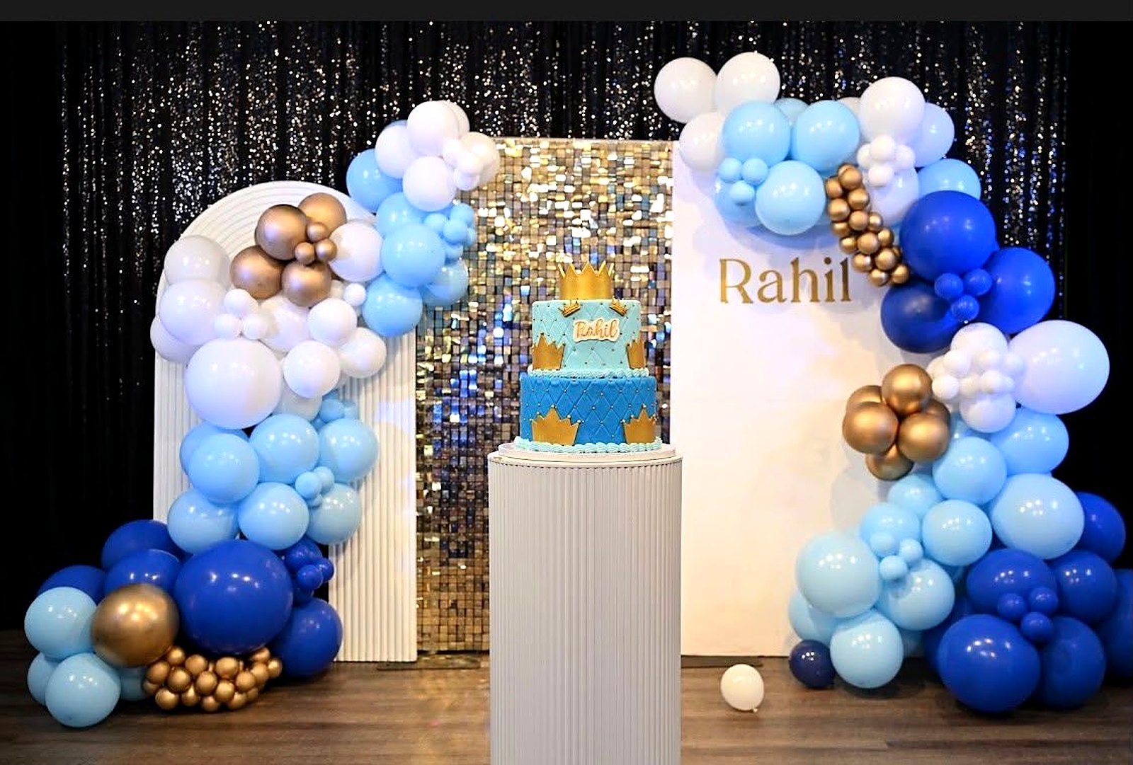 Royal-blue birthday backdrop – blue, white and gold balloon garland, gold sequin wall and a two-tier crown cake
