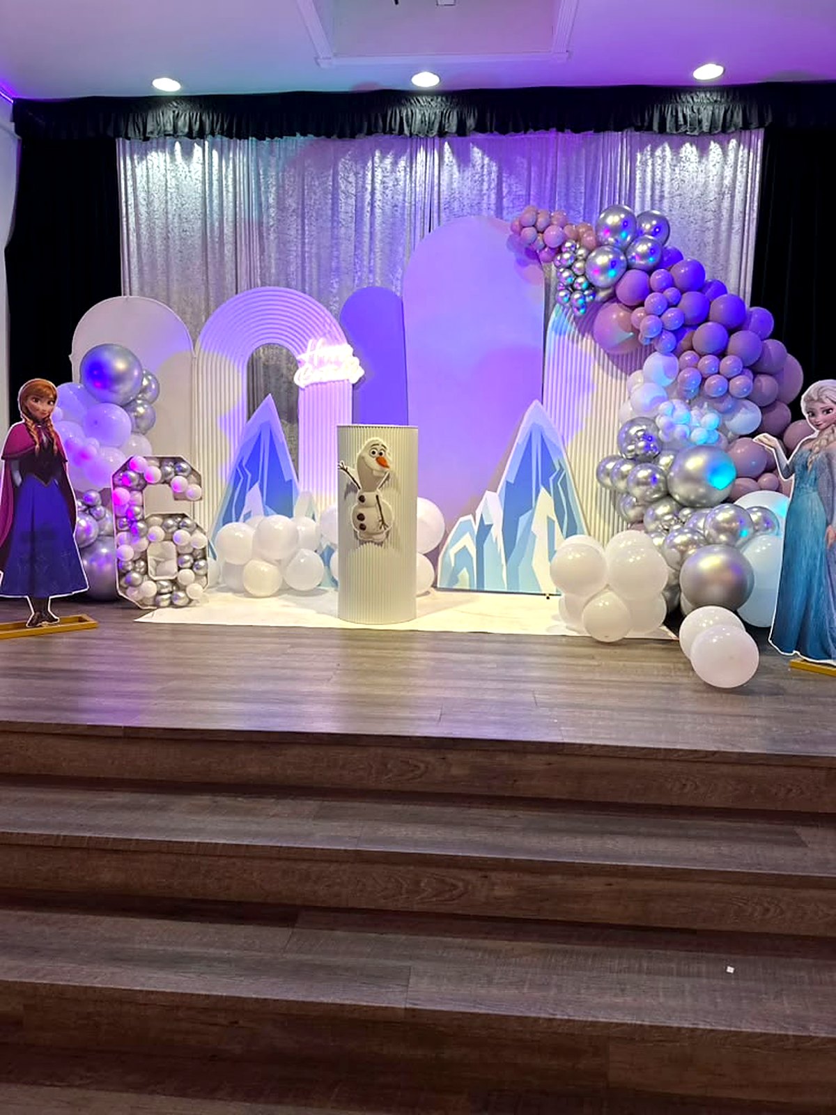 Winter princess birthday stage – icy mountain cutouts, lilac and silver balloons and a “Happy Birthday” neon