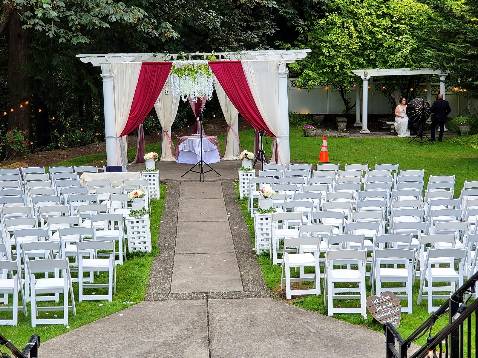 Garden ceremony set-up – pergola in burgundy and ivory drapes with white wisteria, rows of white chairs