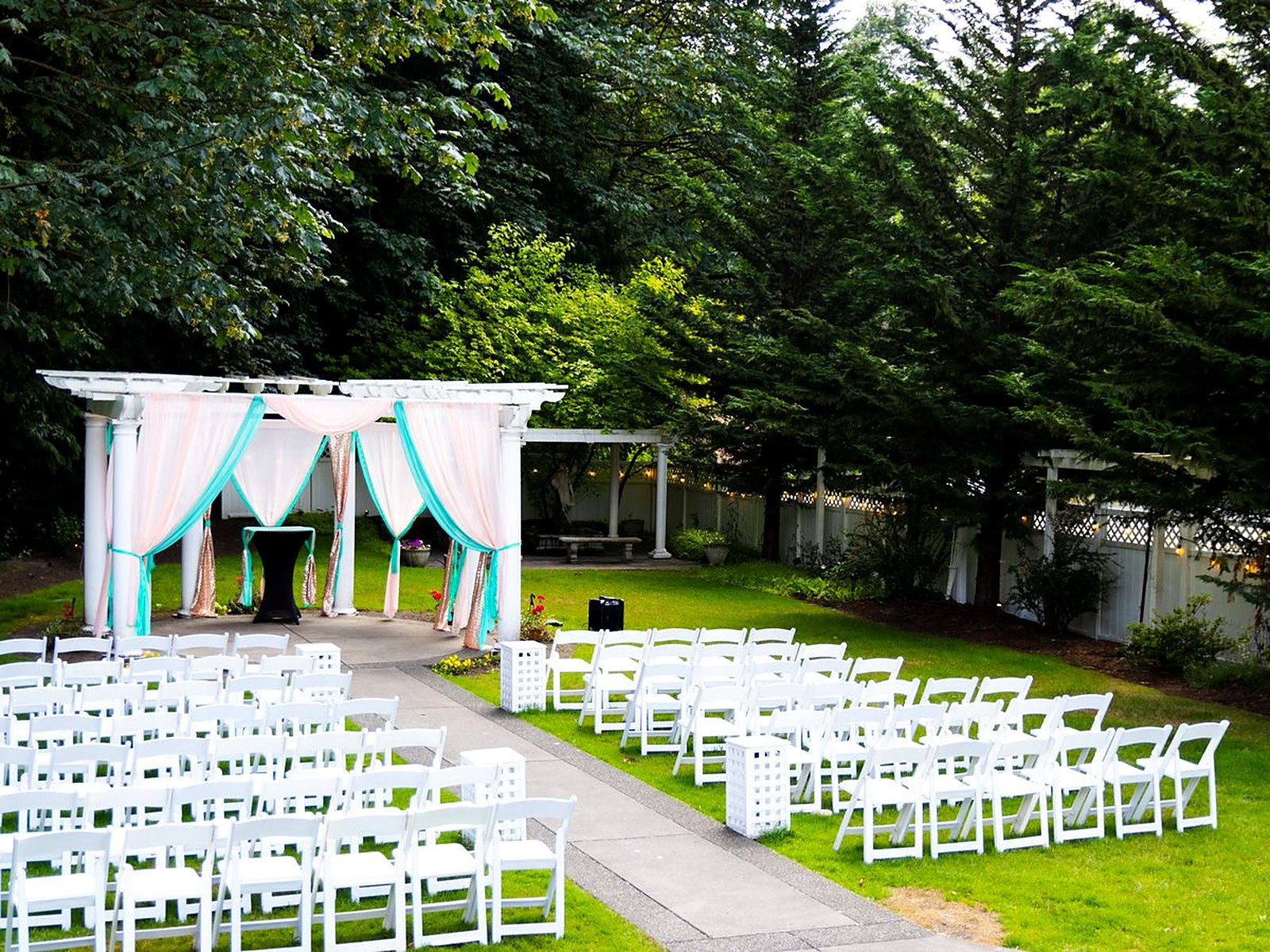 Outdoor garden ceremony – draped pergola with white chairs lining the aisle among the evergreens
