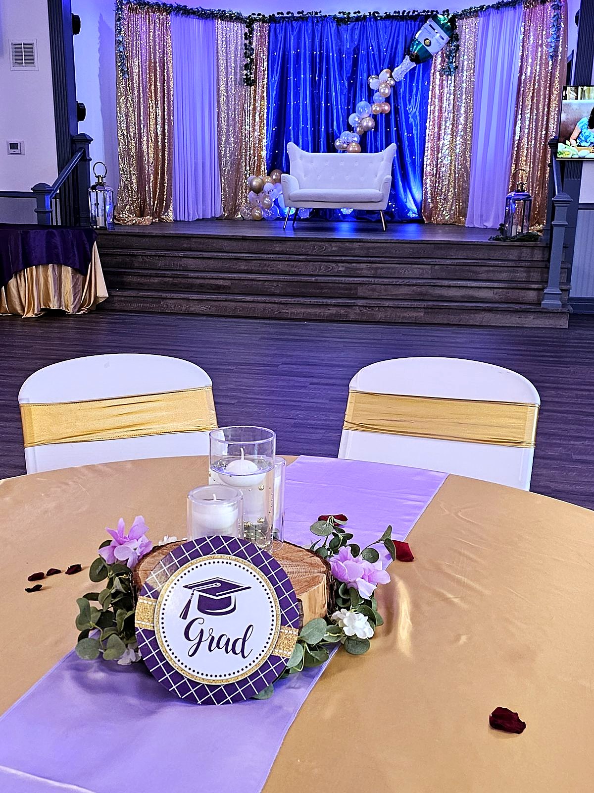 Graduation party table – gold linen, lavender runner and a "Grad" centerpiece, with a blue and gold sequin stage behind