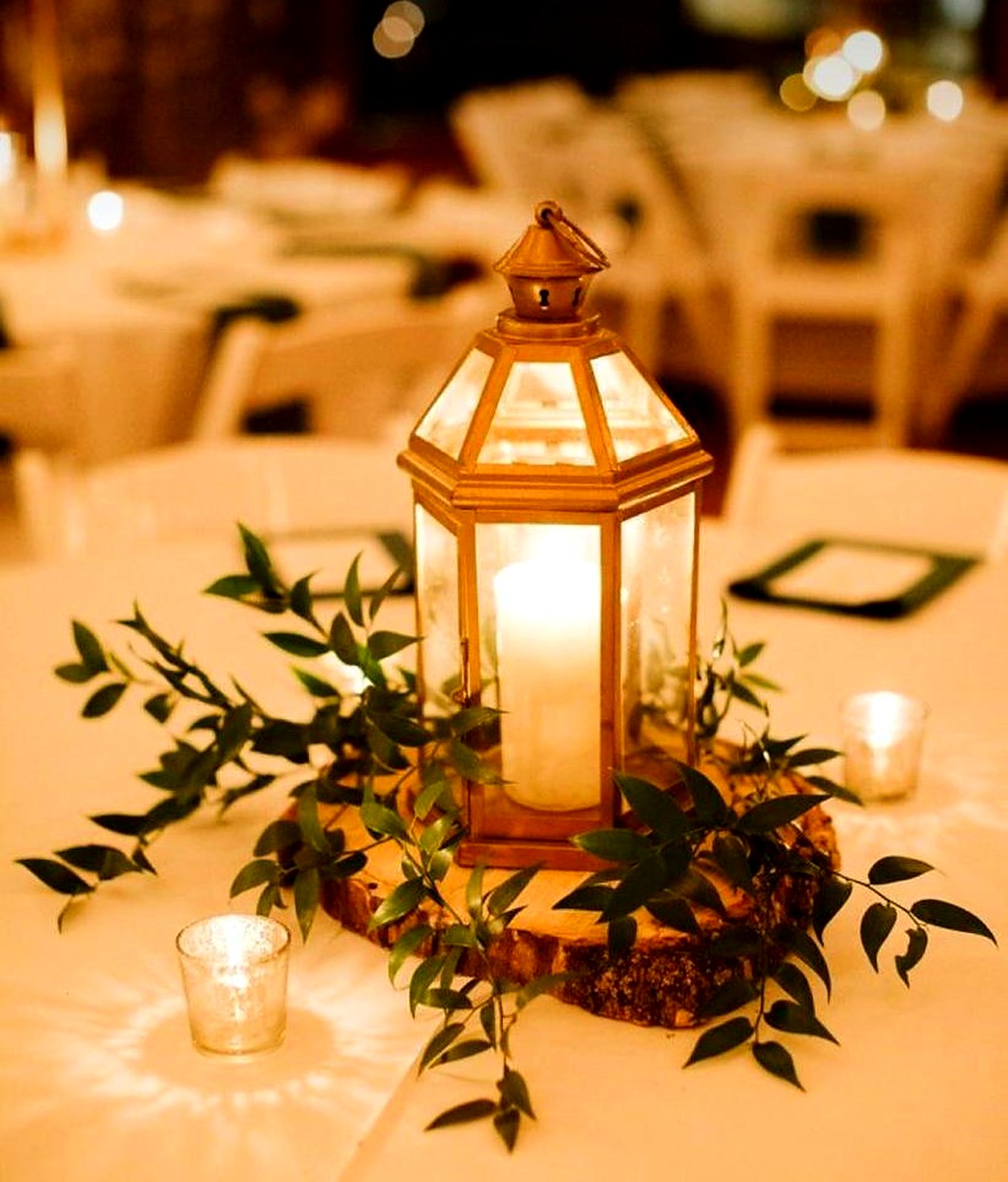 Candlelit gold lantern centerpiece on a wood slice with fresh greenery and votives
