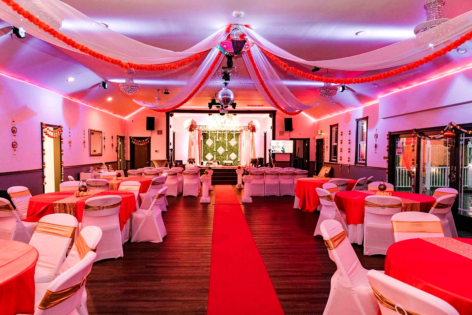 Main hall dressed for an Indian wedding – marigold garlands and white drapes across the ceiling, red tables and a red carpet aisle to the mandap