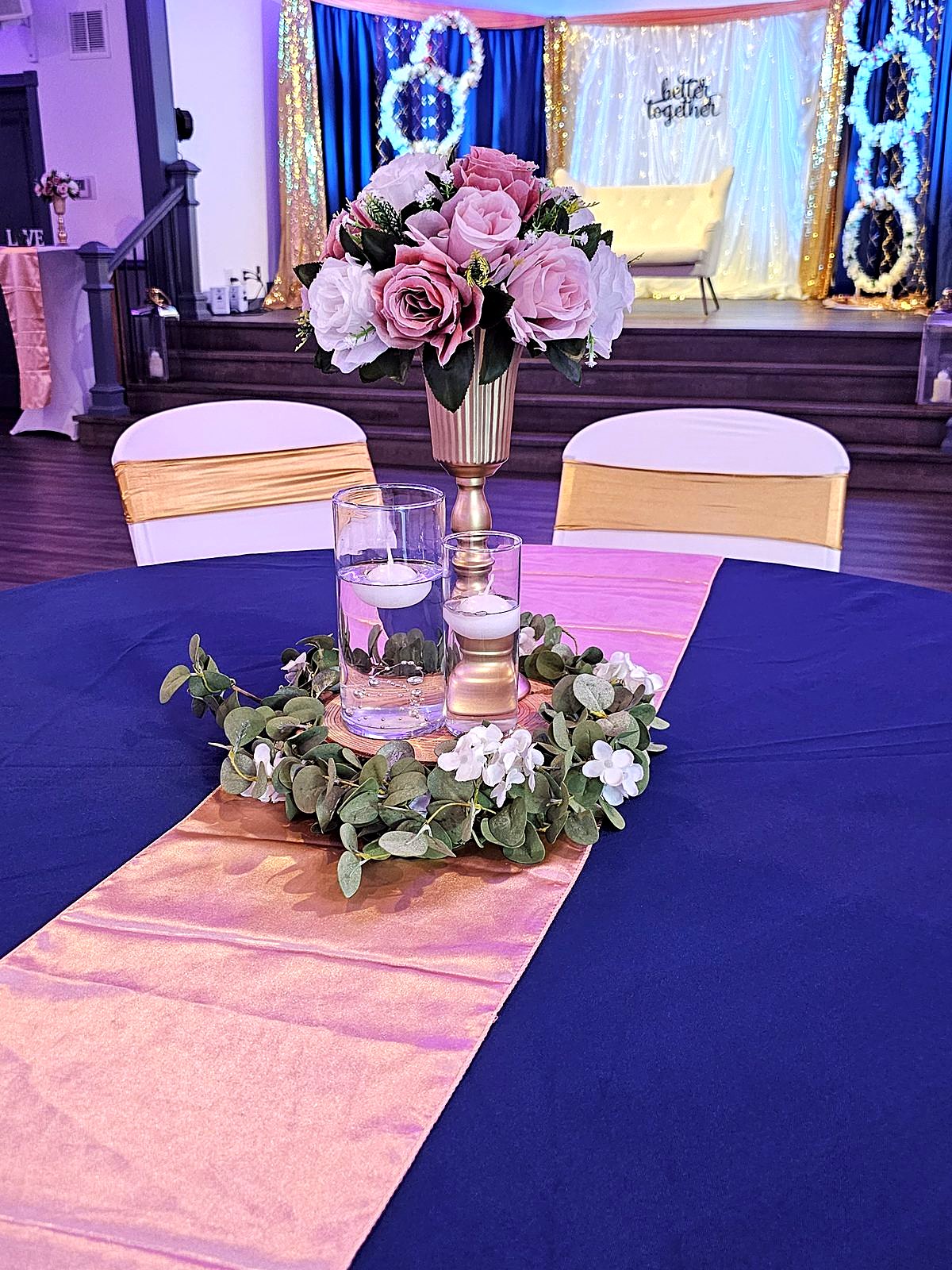 Navy table with a gold vase of blush roses, floating candles and a eucalyptus wreath, facing the "better together" stage