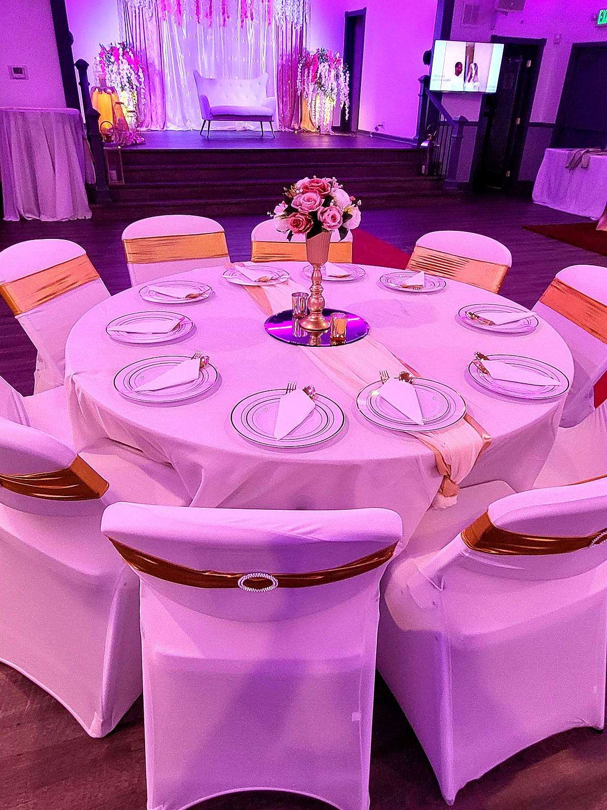 Reception table in the main hall – white linens, gold chair sashes and a rose centerpiece, with the floral loveseat stage behind