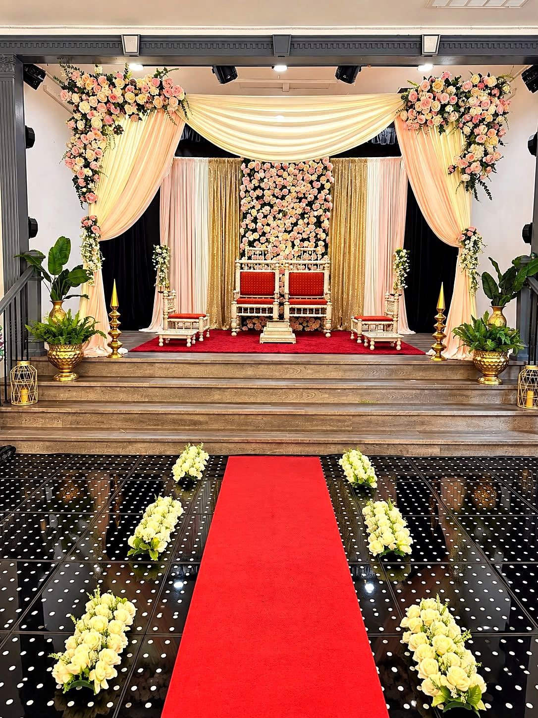 Indoor mandap on the stage – peach and ivory drapes, rose-covered corners, a pink rose wall and carved ivory chairs, with a red carpet aisle across the LED dance floor