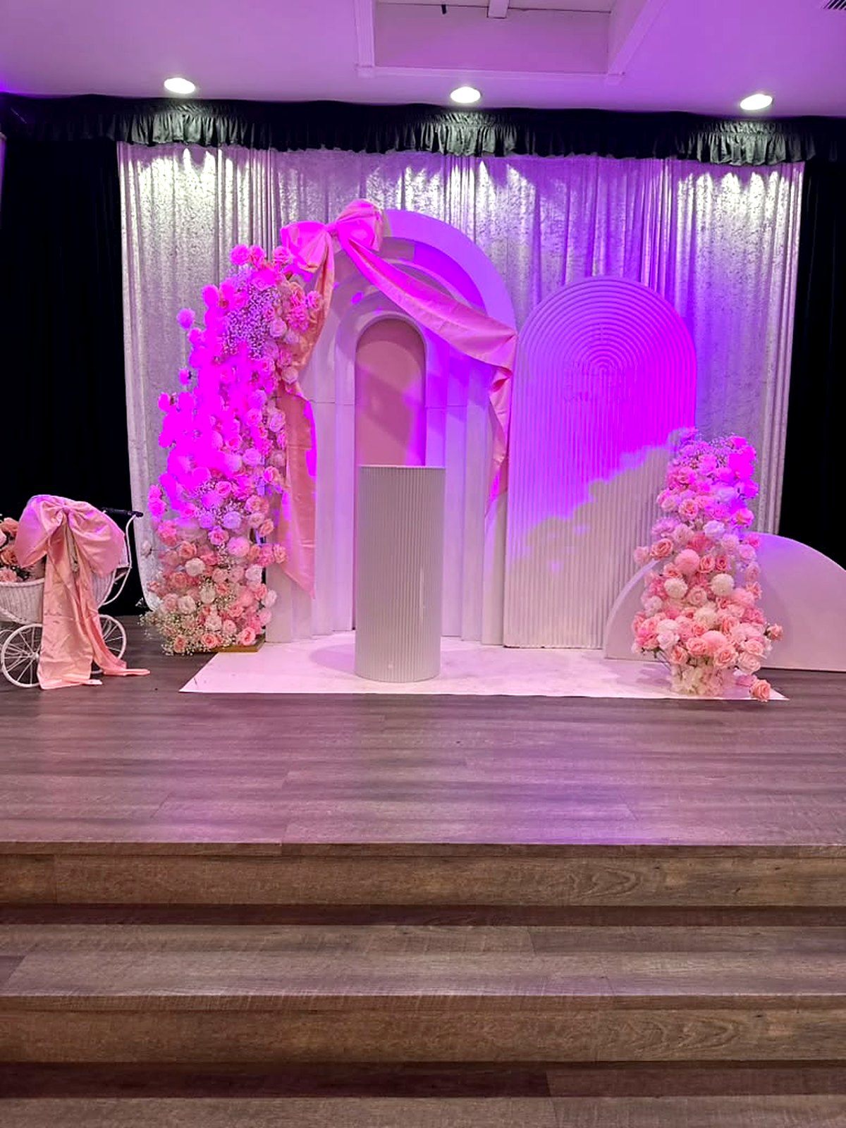 Pink floral baby shower stage – cascading rose columns, satin bow arch and a vintage pram