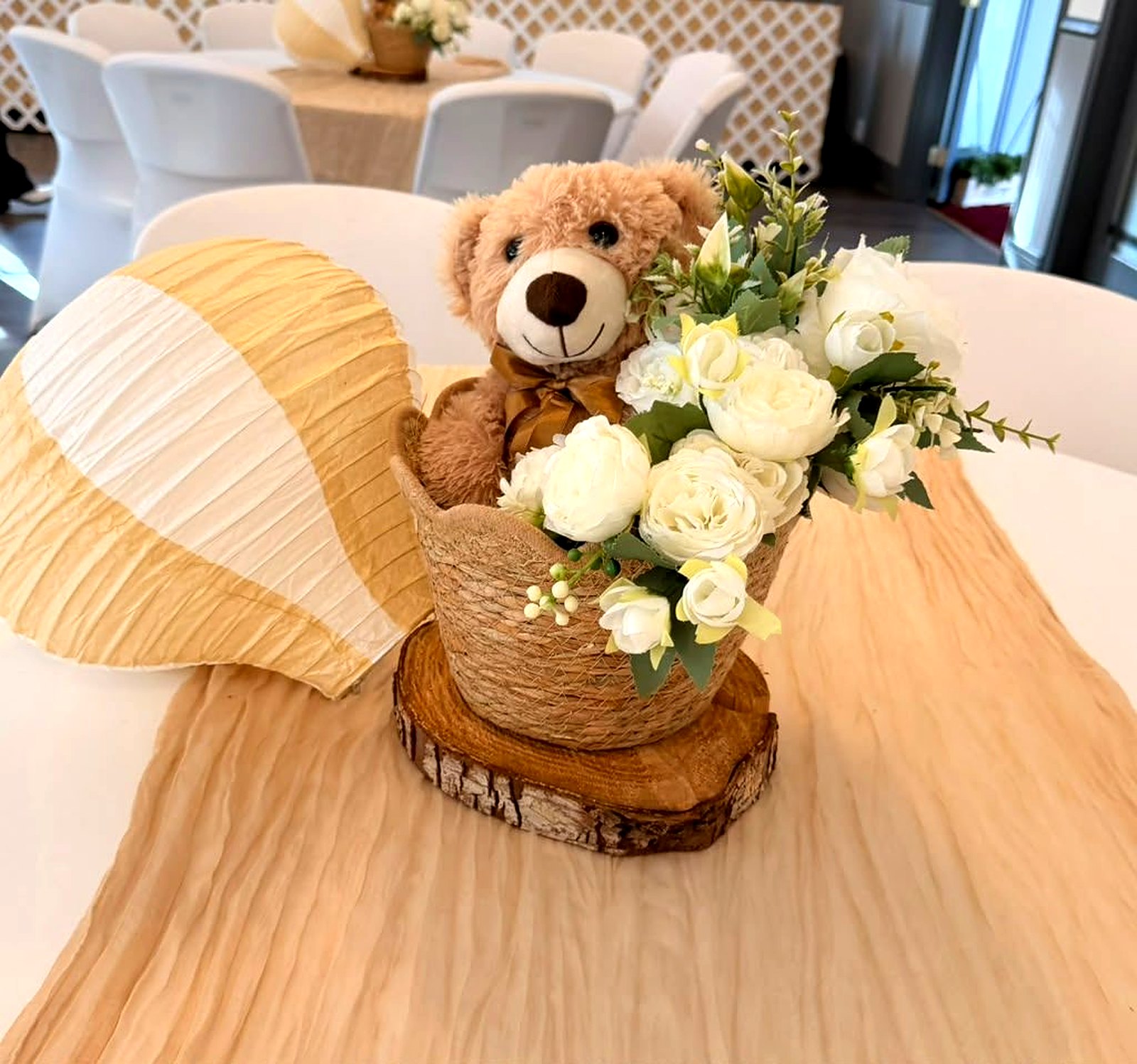 Teddy bear centerpiece in a woven basket with ivory roses on a wood slice, beside a paper hot-air balloon