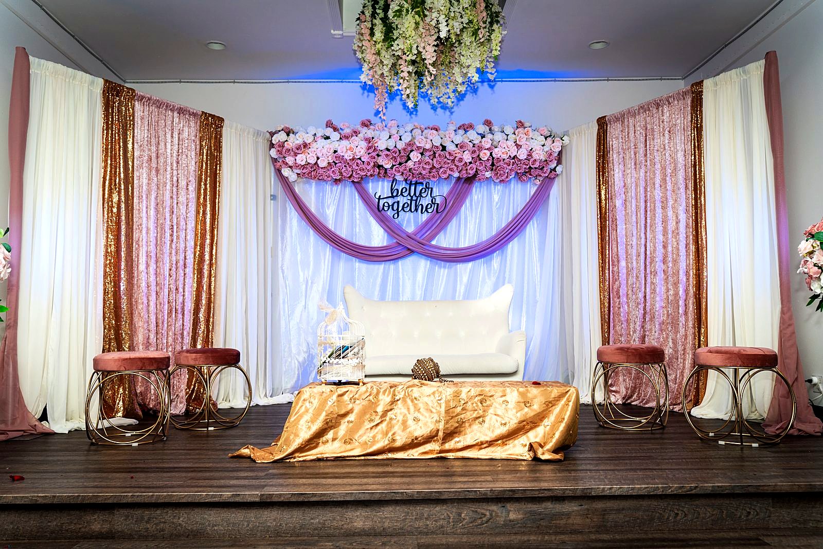 Stage with blush and gold drapes, rose garland and a white loveseat under a "better together" sign