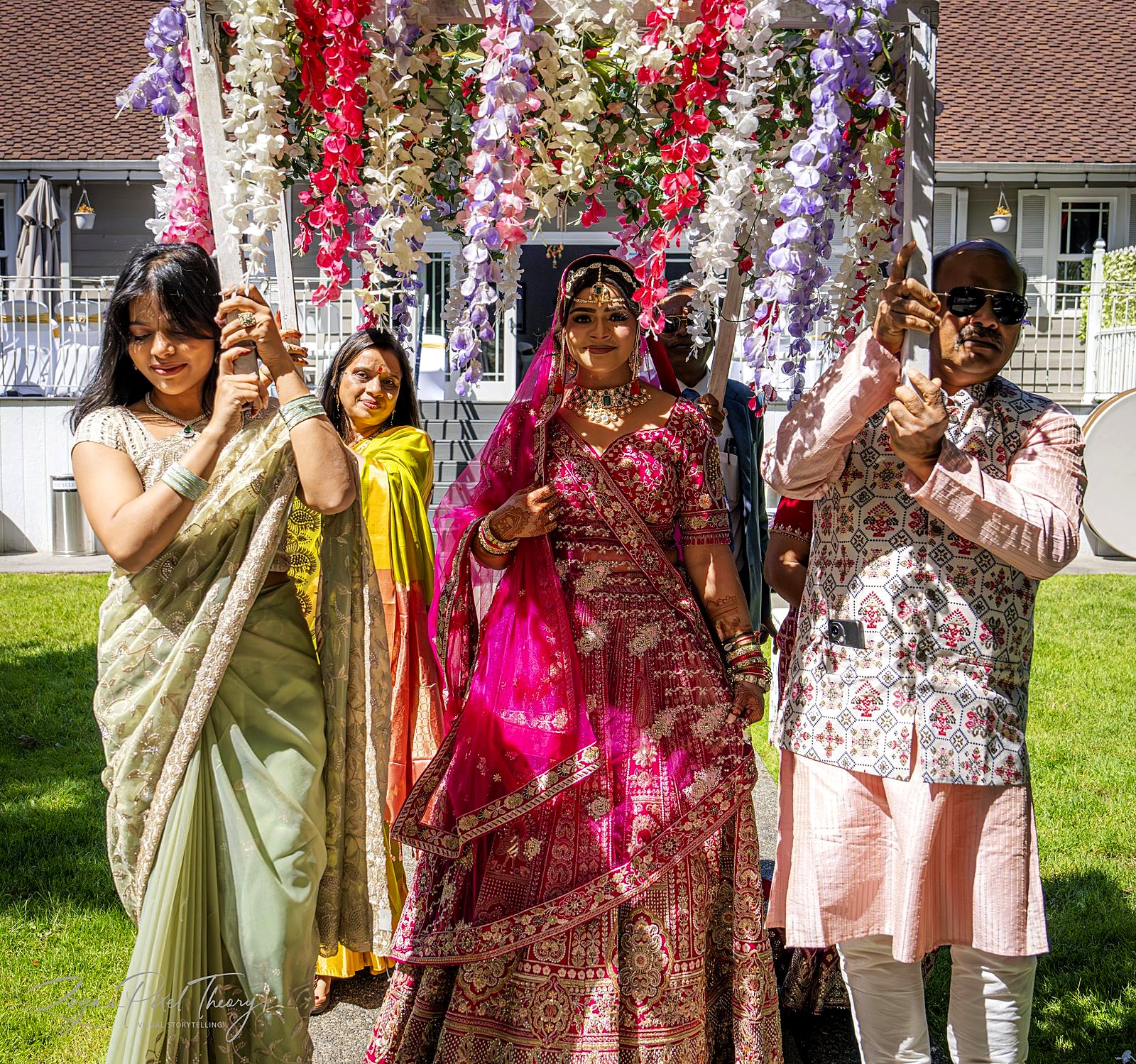 Bride entering under a phoolon ki chadar – a floral canopy of wisteria carried by her family across the lawn
