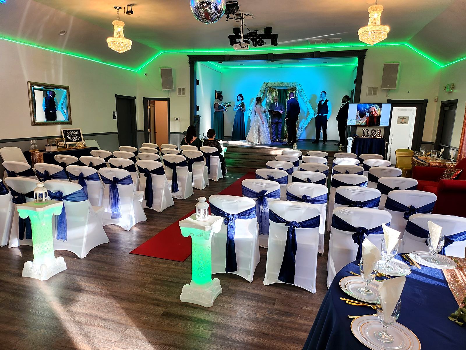 Indoor ceremony on the main stage – white chairs with navy sashes, red aisle runner and glowing pillar lanterns