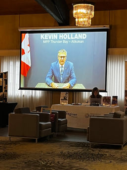 A photo of a screen projector showing a caucasian man in a suit, Kevin Holland, speaking at the conference.