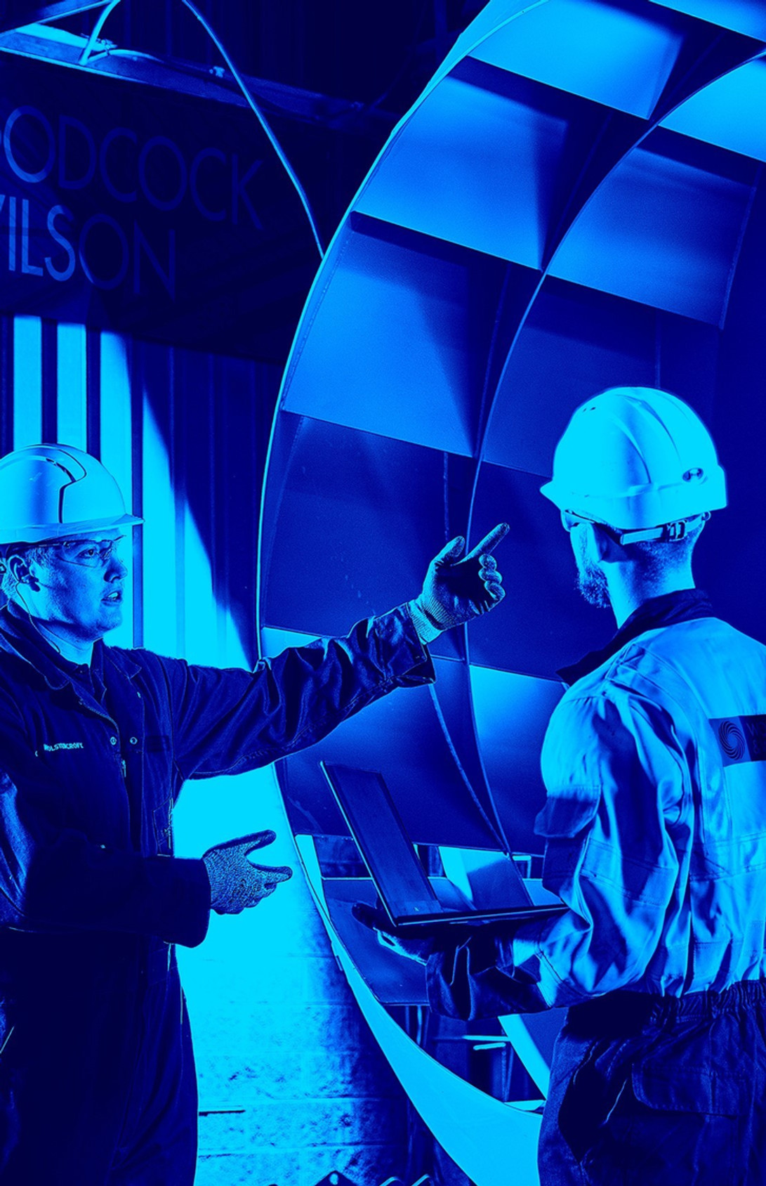 Two hard-hatted workers inspect a large blue industrial fan in a workshop; Woodcock & Wilson sign glows in the background.