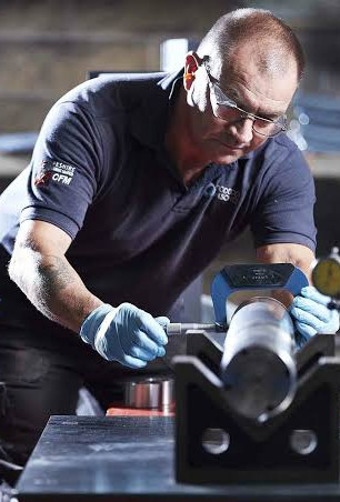 Woodcock and Wilson technician measures a metal cylinder with calipers in a workshop, focused at a workbench.