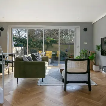 A living room featuring a velet green sofa, chevron wooden flooring, and a view of a garden through large glass doors.