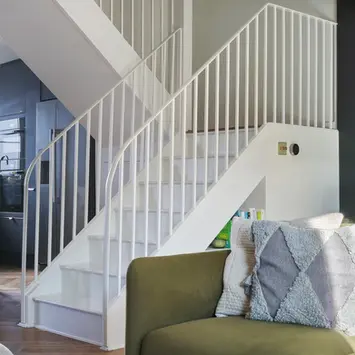 White timber painted stairs winding upwards.  White painted slim metal handrial and balustrade. Green sofa in the foreground with grey cushion. 