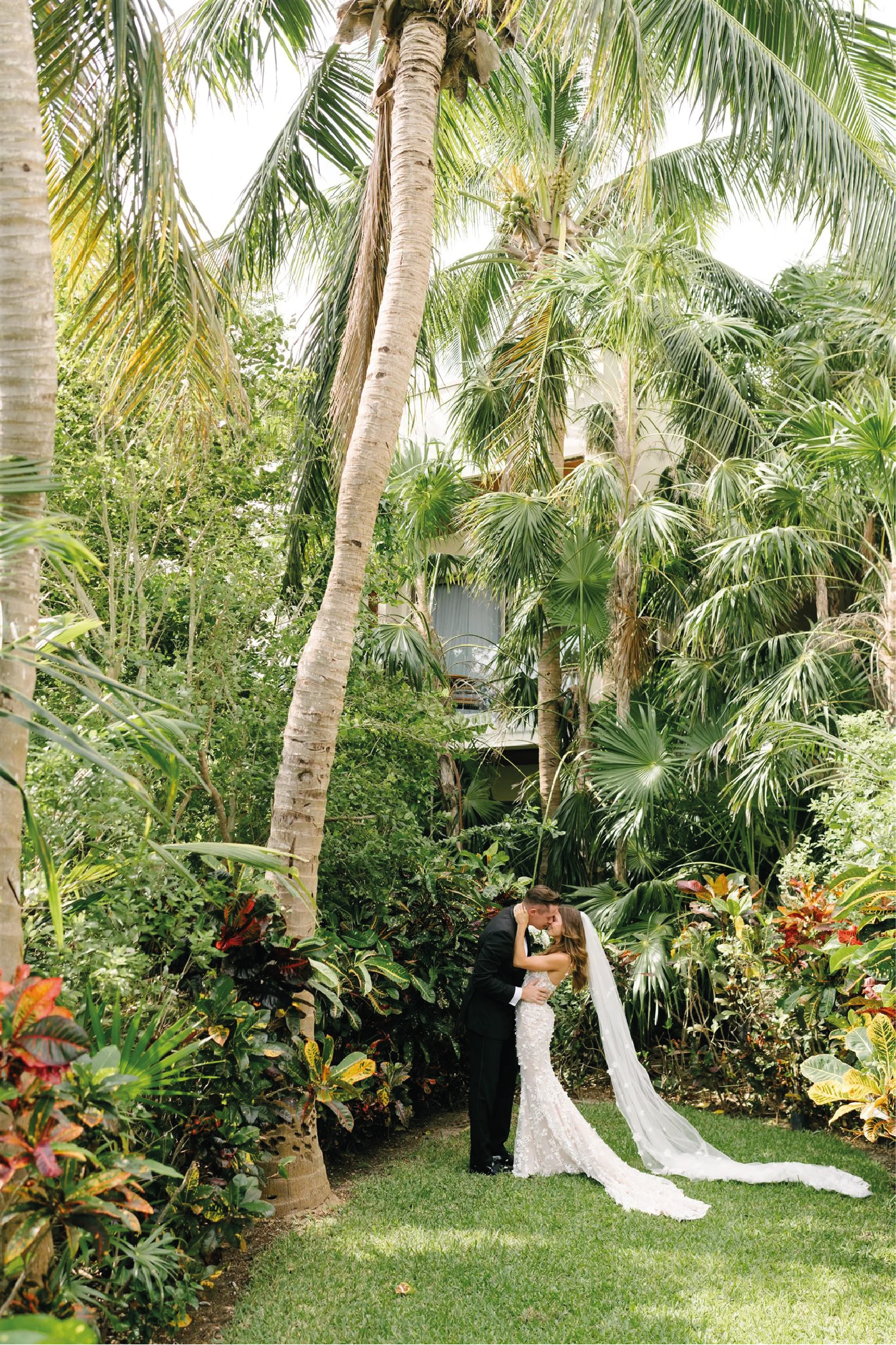 Sam and Matt share a kiss during their outdoor wedding ceremony on the white aisle at Rosewood Mayakoba’s Laguna Lawn in the Riviera Maya. Guests stand in applauding applause, surrounded by elegant white floral arrangements and a backdrop of dense tropical palm trees. Design and production by Bash House.