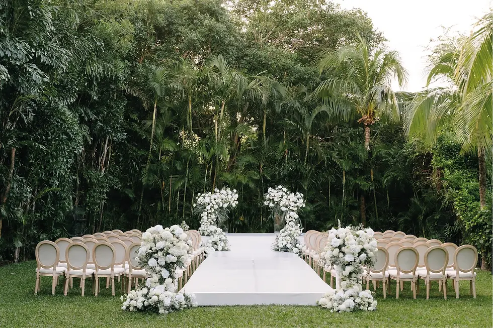 A stunning perspective of the Lagoon Lawn, a premier Rosewood Mayakoba wedding venue, featuring symmetric rows of white Louis XVI-style chairs lining a central white runway. The path leads to a large white floral altar with massive installations, set against a backdrop of dense tropical mangroves and the water's edge. This sophisticated setup for the wedding of Sam & Matt in Cancun Riviera Maya was designed and produced by Bash House.